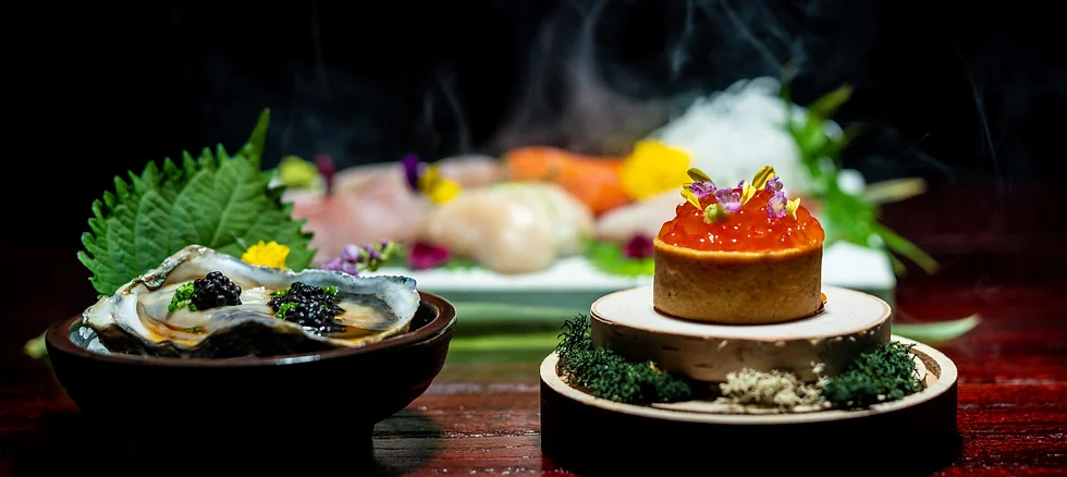Close-up of oysters topped with black caviar in a dark bowl, beside a small tart topped with orange roe and flowers, with a moody, elegant dark-toned presentation