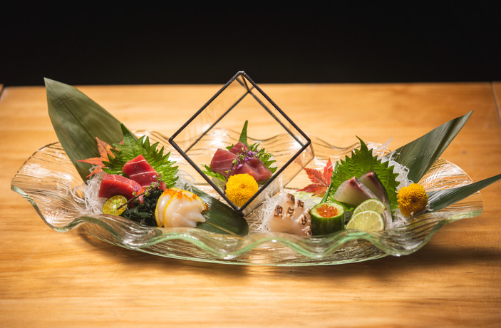Assorted sashimi slices arranged on a wavy glass dish with bamboo leaves, citrus, roe, and flower garnishes, in soft natural light with muted greens and warm orange accents