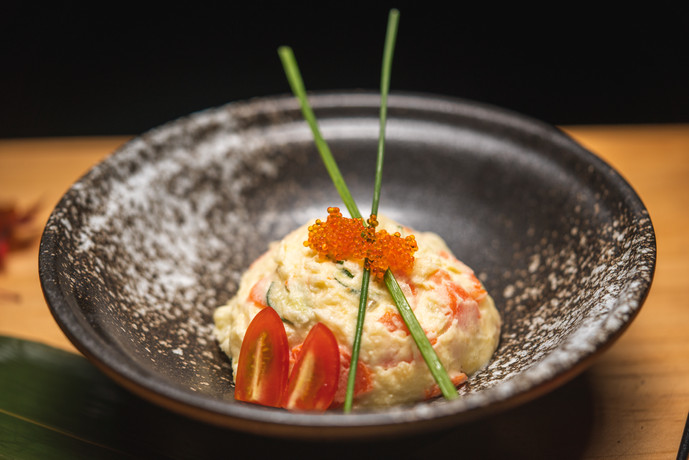 A scoop of creamy salad topped with orange roe and chive sticks, garnished with cherry tomato slices, served in a speckled bowl against a soft-focus warm background