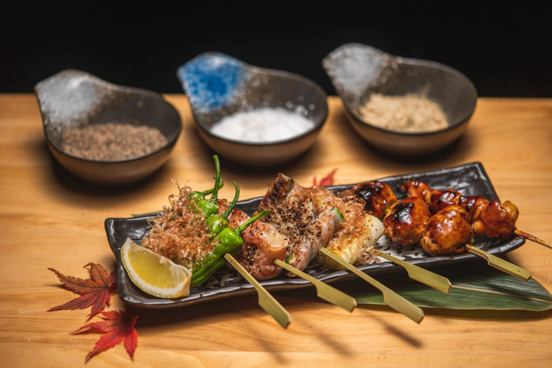 Grilled skewers of meat and vegetables on a dark rectangular plate with lemon wedge and maple leaf garnish, set before a warm, dimly lit dining backdrop