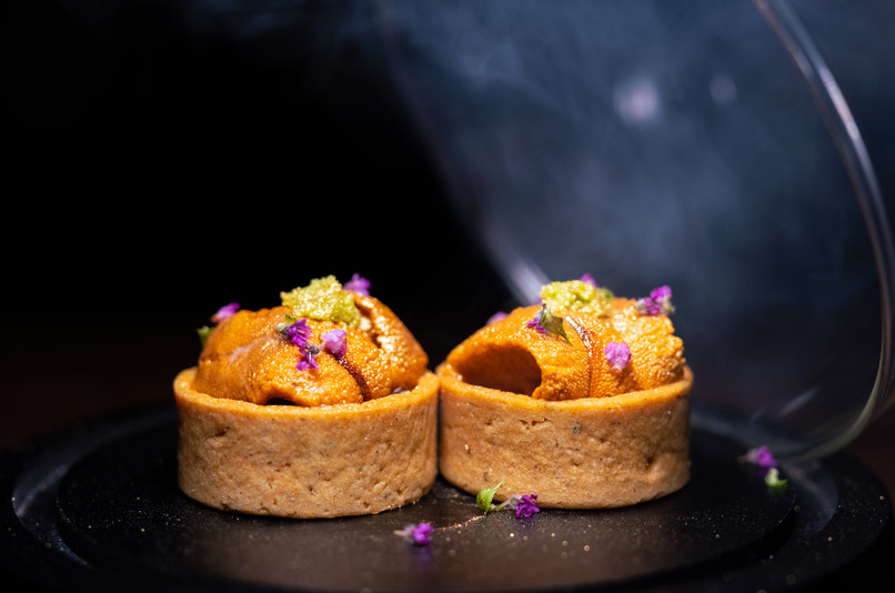 Two small pastry tart shells filled with orange sea urchin, garnished with purple flowers, on a dark plate with smoke effect